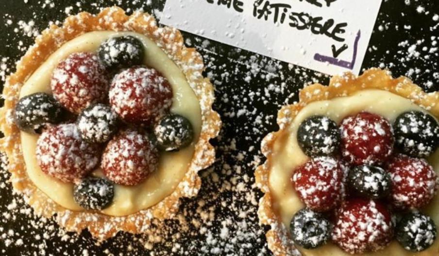 Close-up of a fruit tart topped with blueberries and raspberries, dusted with powdered sugar, next to a handwritten label.