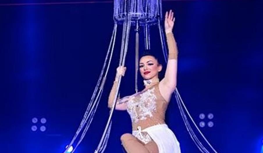 A female performer in a sparkling, sheer costume poses gracefully on a large, hanging chandelier prop with blue stage lights in the background.