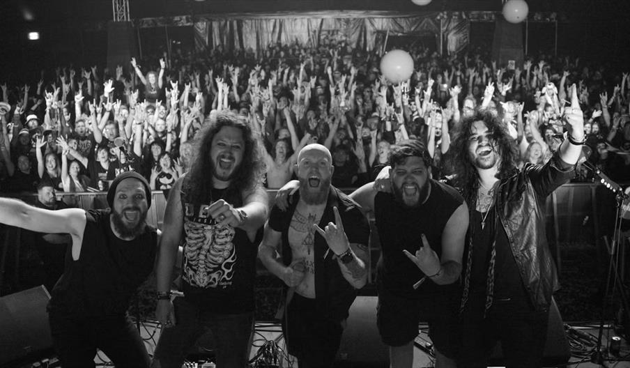A black-and-white photo of five enthusiastic band members posing at the edge of a stage, with a large, cheering crowd behind them raising their hands