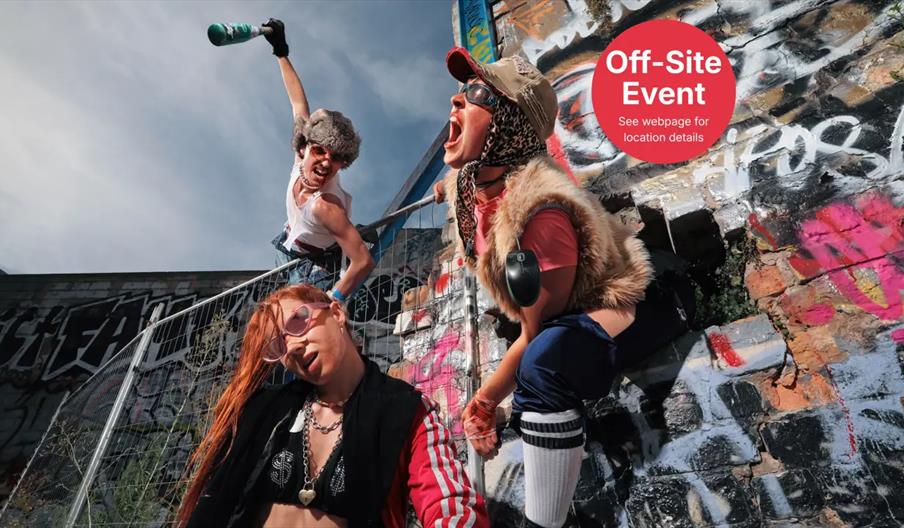 Three people in eclectic, punk-inspired outfits pose energetically against a graffiti-covered wall and fence outdoors. A red circle in the upper right