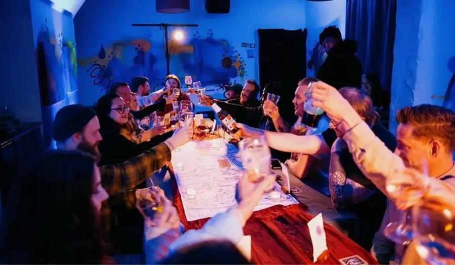 A group of people seated around a long table raise their glasses in a celebratory toast, illuminated by colorful lighting in a lively indoor setting.