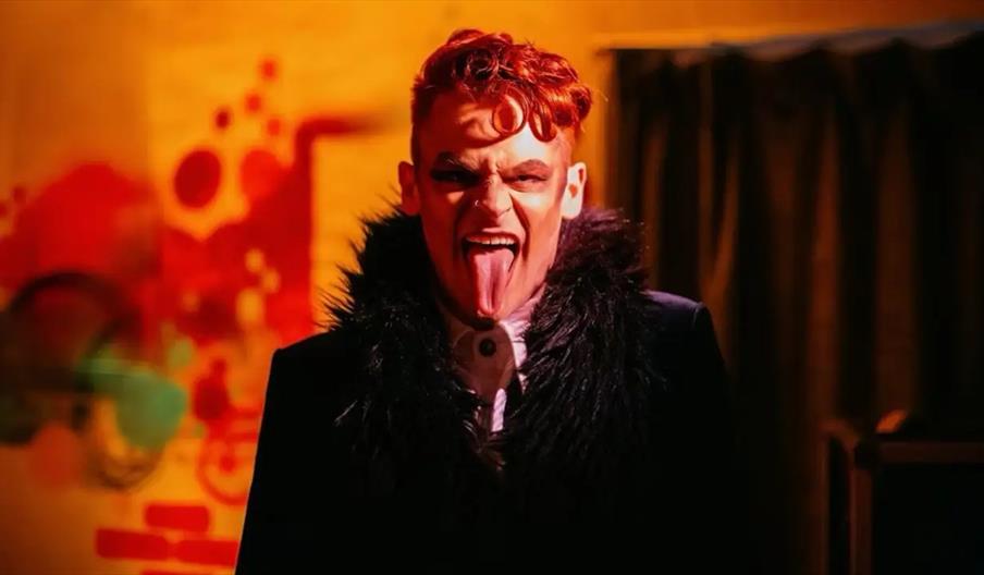 A person with bright red hair and dramatic makeup stands in front of a colorful, abstract background, sticking out their tongue and wearing a dark out