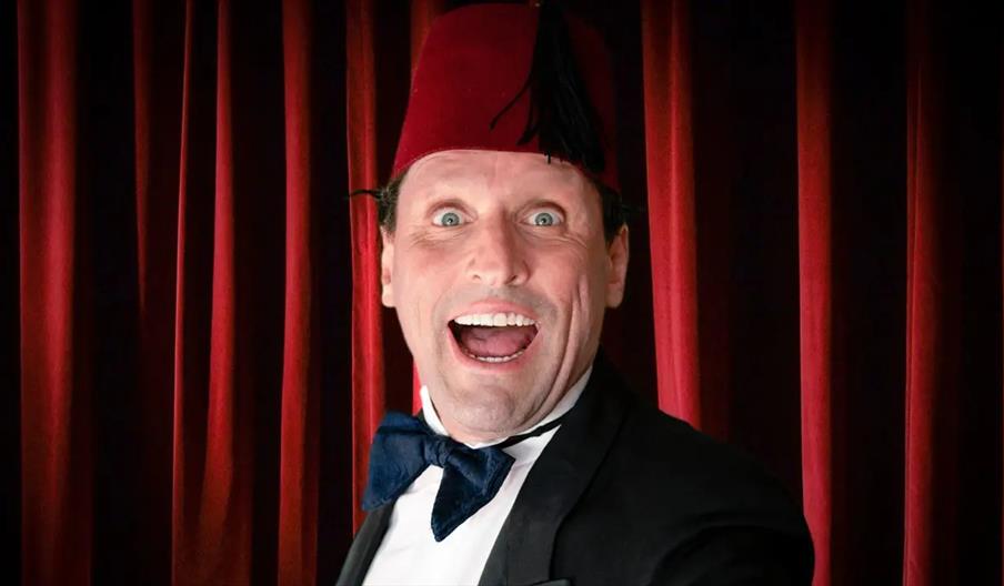 A smiling man wearing a red fez hat, tuxedo, and blue bow tie stands in front of red stage curtains with a wide, excited expression.