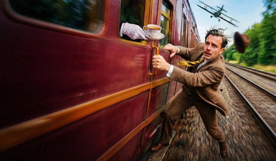 A man in a brown suit clings to the side of a moving maroon train, with his tie and hat flying behind him; a gloved hand offers him a teacup from a tr