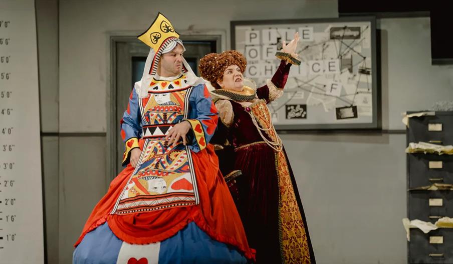 Two actors in elaborate costumes stand on a stage; one is dressed as a playing card queen and the other in a royal gown with a large curly wig, gestur