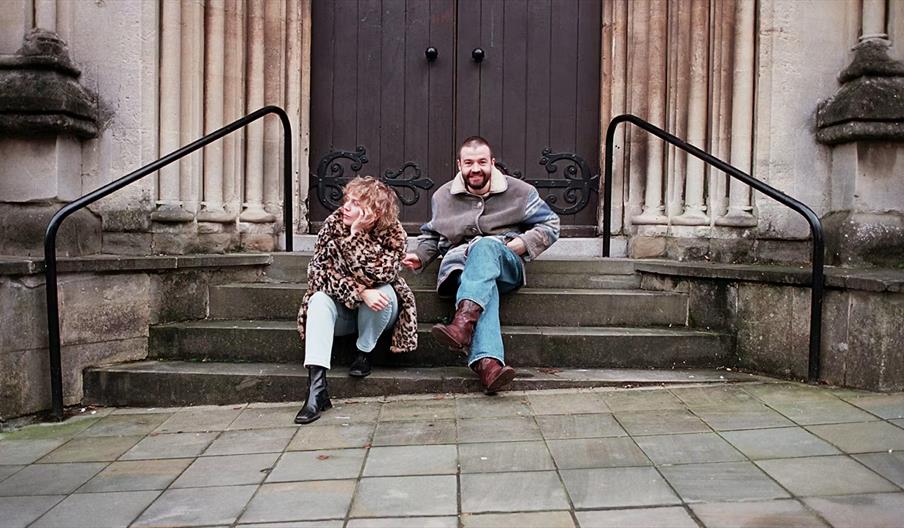 Two people sit on stone steps in front of a large, dark wooden door with ornate metalwork. One person wears a leopard print coat and boots, while the