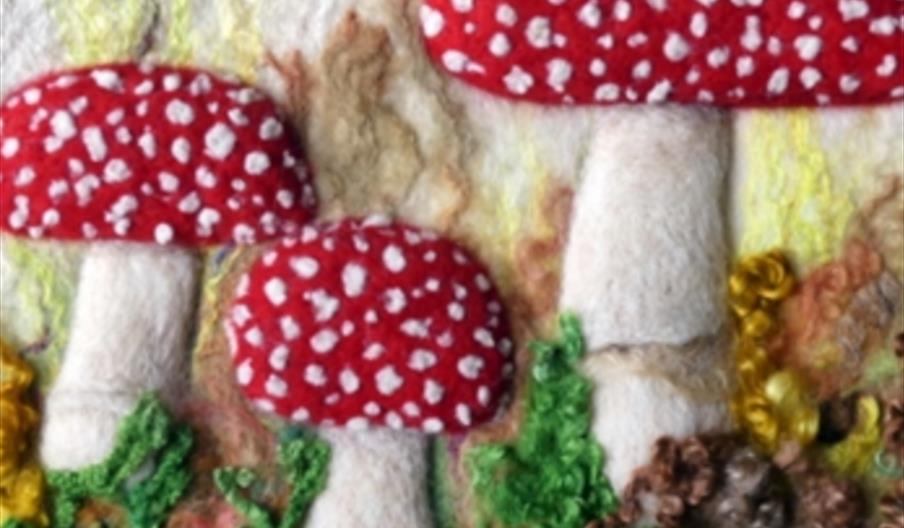 A textile art piece depicting three red mushrooms with white spots, resembling fly agaric, standing among green foliage and brown elements on a textur
