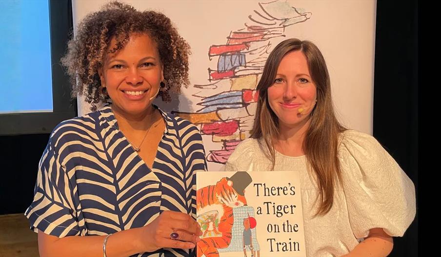 Two women are standing side by side, smiling at the camera. One woman is holding up a children's book titled "There's a Tiger on the Train," and there