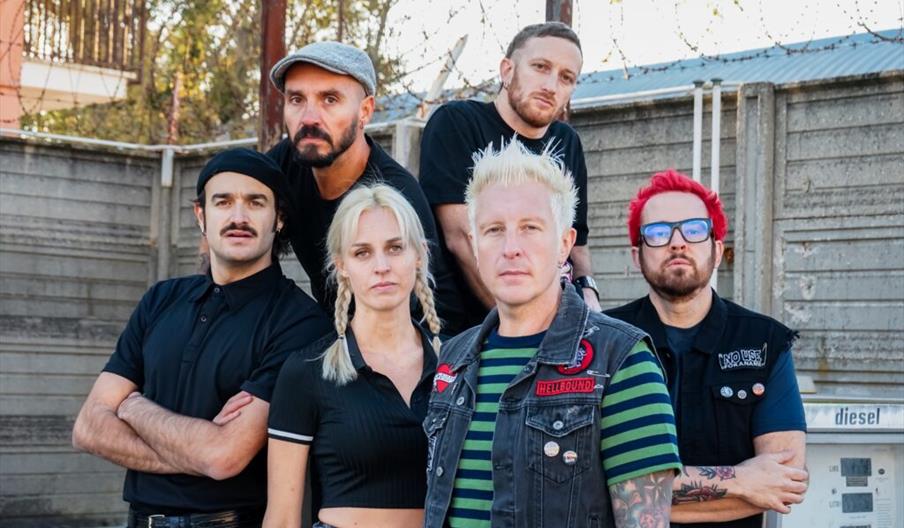 Six people stand together in front of a concrete wall topped with barbed wire, looking seriously at the camera; four men and one woman are in the fron