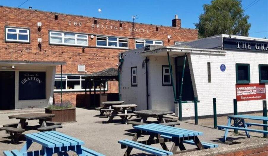 The exterior of The Grafton Pub, with blue and brown picnic benches out front and a planter.