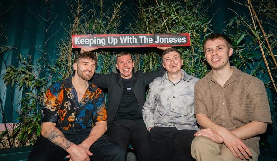 Four smiling young men sit together in front of green foliage, with one holding up a sign above their heads that reads "Keeping Up With The Joneses."