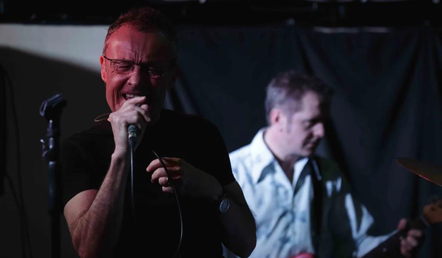 A man in the foreground is passionately singing into a microphone with his eyes closed, while another man in the background plays an electric guitar.