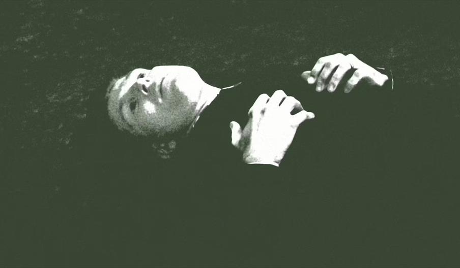 A person lies on their back with hands clasped on their chest, looking upward. The image has a dark, high-contrast, monochromatic filter, giving it a