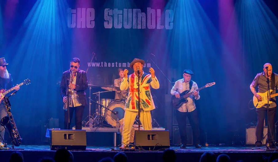 A six-member band performs on stage under colorful lighting, with the lead singer at the center wearing a vibrant jacket and hat, flanked by musicians