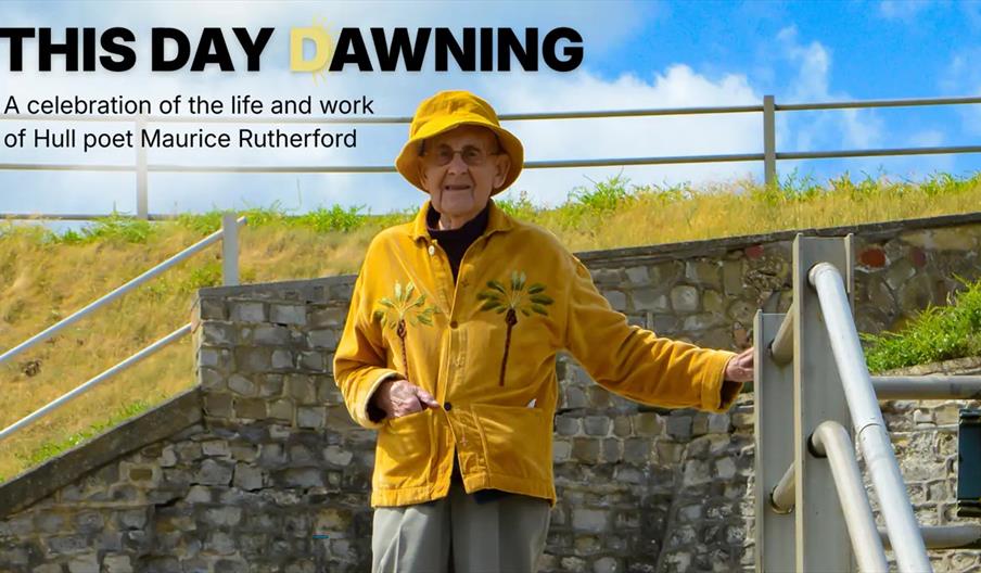 An elderly man in a yellow jacket and matching hat stands outdoors near stone steps and a railing, with grassy hills and a blue sky in the background.