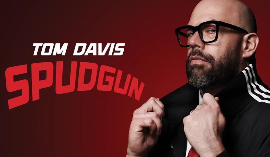 A man with a bald head, beard, and glasses adjusts the collar of his black jacket with white stripes on the sleeves. The background is a red gradient,