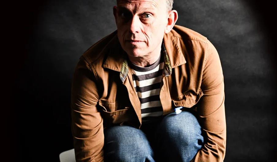 Tom Hingley with short hair, wearing a brown jacket over a black and white striped shirt and jeans, is crouched on a white chair against a dark backgr