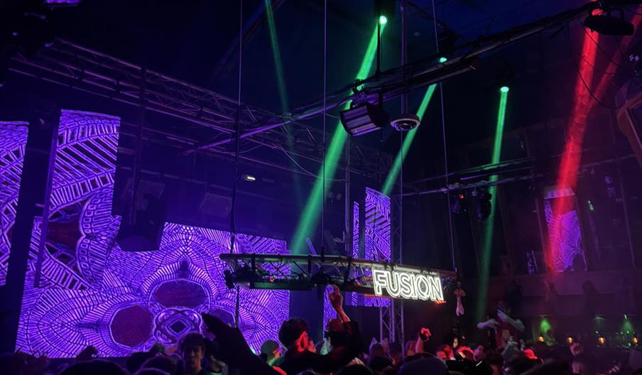 A crowded nightclub scene with people dancing under colorful green and red spotlights, a large LED screen displaying vibrant purple patterns in the ba