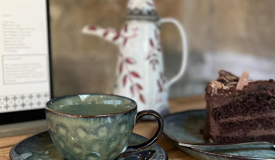 A cup of tea and a slice of chocolate cake on a plate on a wooden table with a menu in the background.
