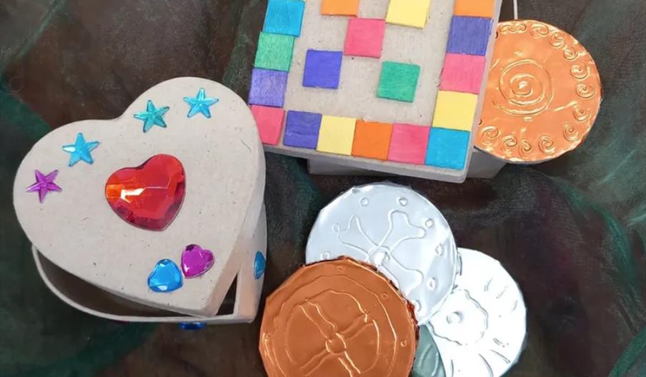 Two small craft boxes, one heart-shaped and decorated with colorful gems, and one square-shaped with a mosaic of colored squares on the lid, are shown