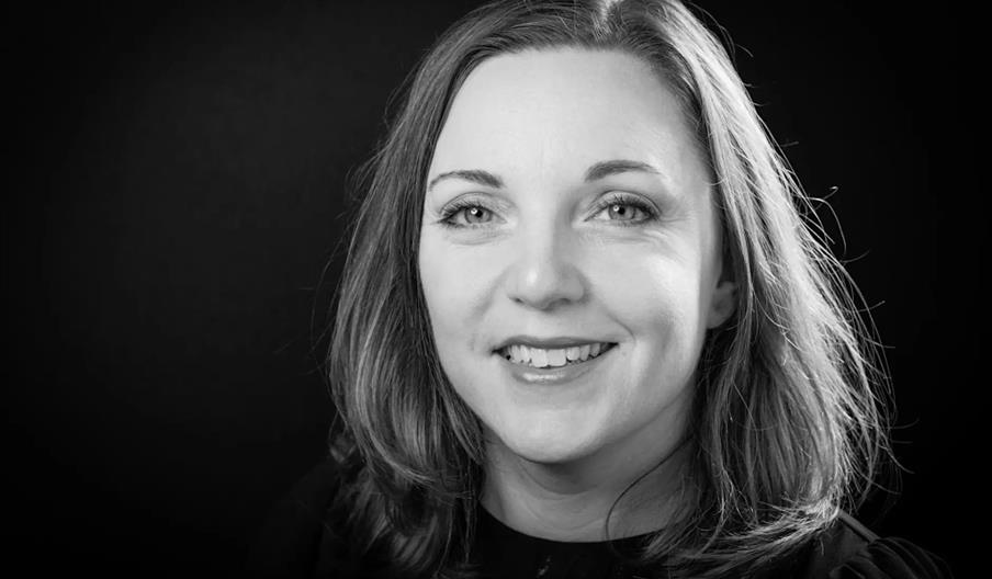 Black and white portrait of a woman with shoulder-length hair, smiling and looking at the camera, set against a dark background.