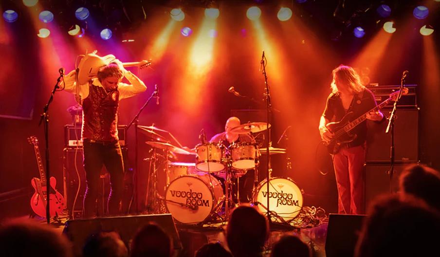 A three-piece band performs on stage under dramatic lighting, with the guitarist holding a white electric guitar over his shoulders, the drummer playi