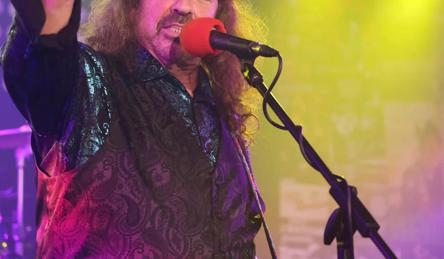 A male musician wearing a black patterned shirt and hat plays electric guitar and sings into a microphone on stage, with yellow stage lights shining i
