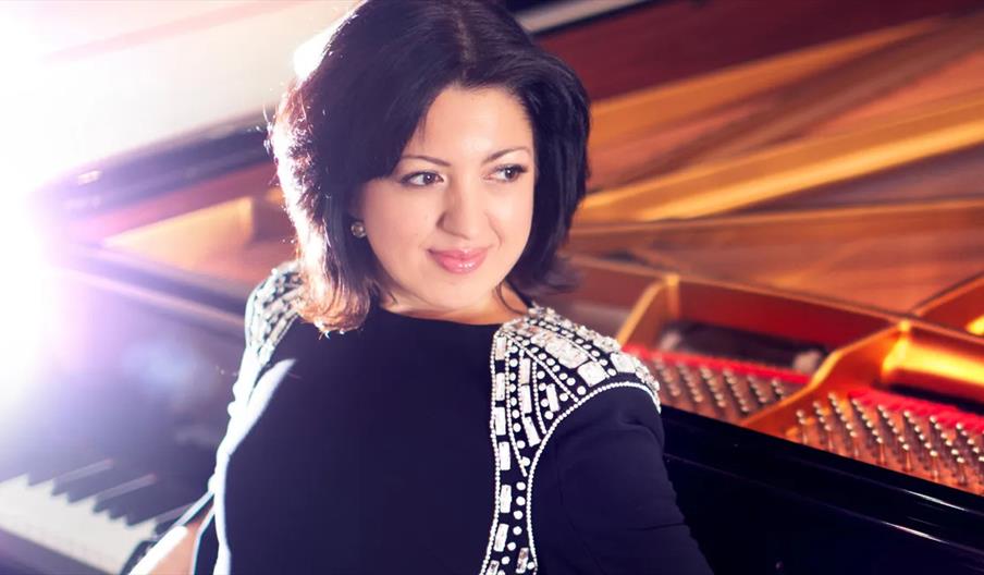A woman with dark hair and a black dress decorated with shiny embellishments poses next to an open grand piano, smiling slightly and looking to the si