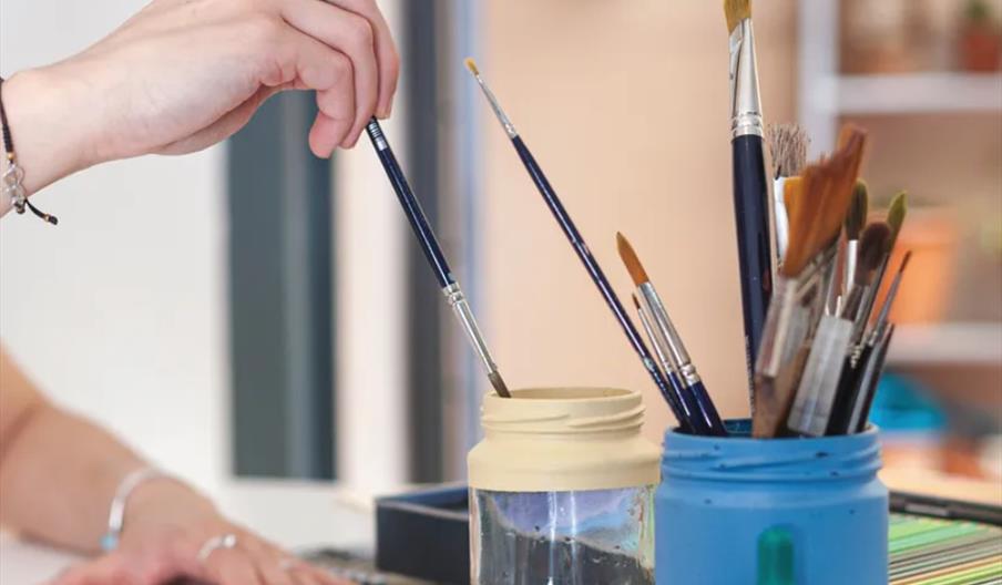 A person dips a paintbrush into a jar of water, with several other paintbrushes standing upright in jars nearby on a table.