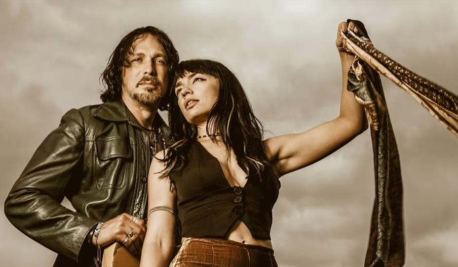 A man and woman stand closely together in front of a cloudy sky, both looking confidently past the camera; the man wears a leather jacket, and the wom