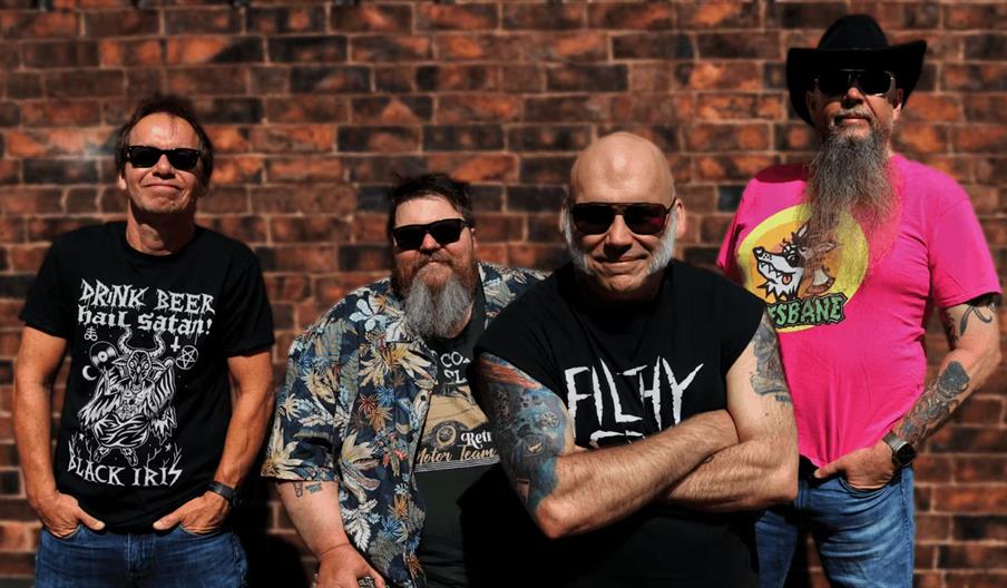 Four men stand in front of a brick wall, all wearing sunglasses and casual clothing, including t-shirts with various graphics and tattoos visible on t
