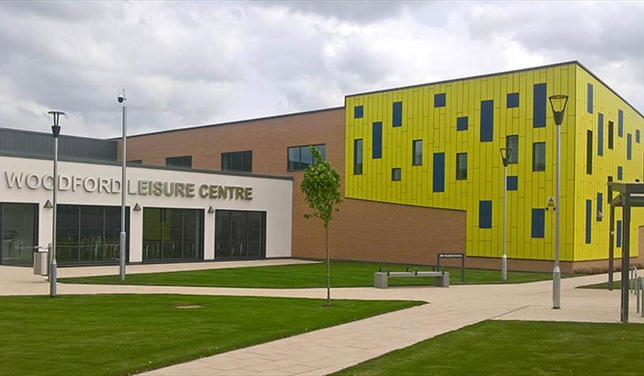 Modern building labeled "Woodford Leisure Centre" with large glass doors, a white and tan facade, and an adjoining bright yellow wing featuring scatte