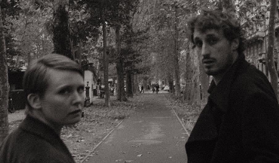 Two people, a woman and a man, stand on a tree-lined path looking back toward the camera in a black-and-white, slightly blurry image. The path is cove