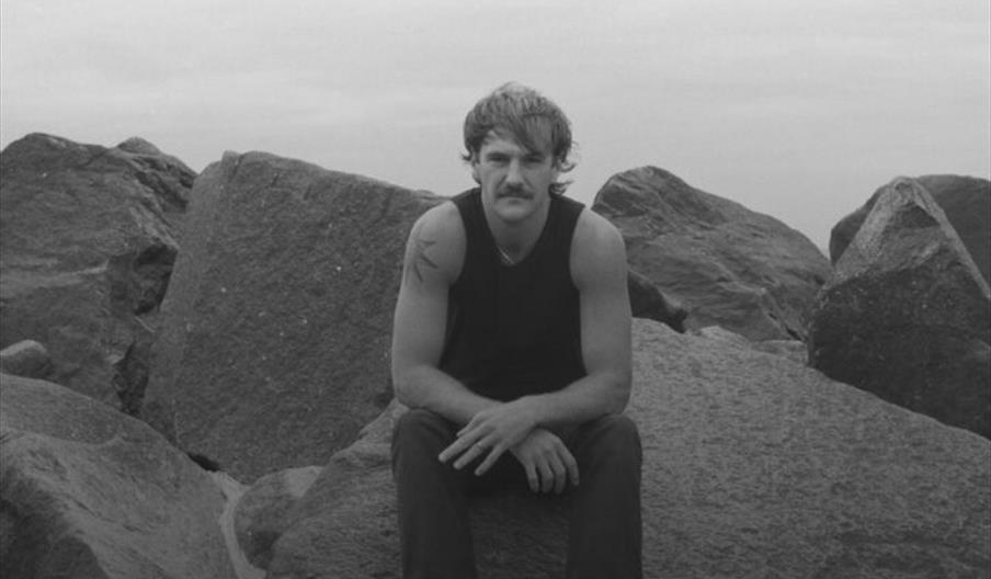 A black and white promotional poster for a music event features a man with a mustache sitting on rocks in the center. Large red text at the top reads