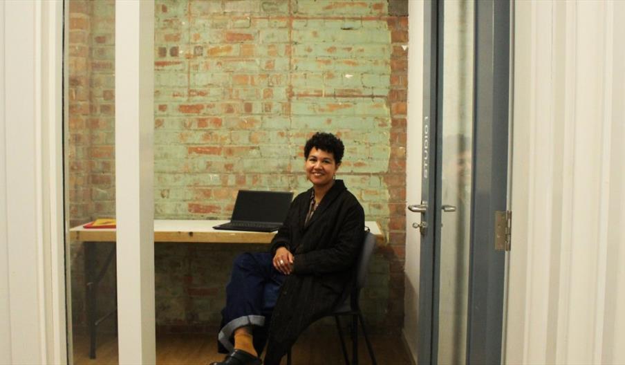 A person sits on a chair at a desk in a small office with exposed brick walls, smiling at the camera. There is a closed laptop on the desk and the roo