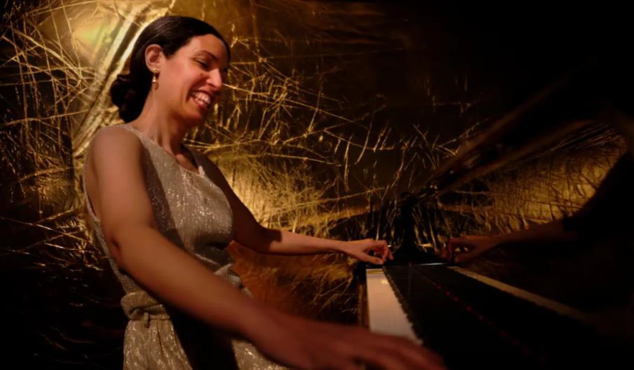 A woman in a sparkly dress smiles joyfully while playing a grand piano, with warm golden lighting and an abstract textured backdrop behind her.
