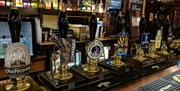 A row of traditional beer taps with various ale labels in a warmly lit pub, with shelves of liquor bottles and glassware in the background. The taps f
