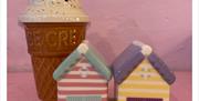 A ceramic ice cream cone with a swirl of white ice cream and colourful sprinkles stands behind two small ceramic beach huts, one pink and blue striped