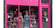A brightly coloured storefront with bold pink framing and large glass windows displays two mannequins in dresses and several eclectic items on shelves