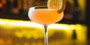 Pink cocktail in a coupe glass with a citrus garnish on a bar counter.