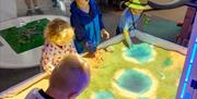 3 children play in a sandpit with a projection mapped over the top, giving different heights different colours.