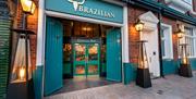 The image shows the entrance to a restaurant called "The Brazilian Churrascaria & Bar," featuring teal double doors, a sign with a bull skull, and out