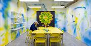 A person in a cap sits at a yellow table with yellow chairs surrounding it inside a gallery with white walls that have been covered in yellow, green a