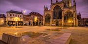 A nightime image of Hull Minster, a large gothic style Minster with stained glass windows and spires. In front there is a wide open paved area sports