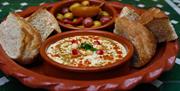 A clay platter holds pieces of rustic bread, a bowl of hummus garnished with spices, chickpeas, and pomegranate seeds, and a small dish of mixed olive
