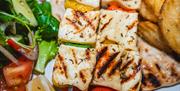 Grilled halloumi cheese skewers with vegetables are served alongside potato wedges, a mixed salad with tomatoes and greens, pita bread, and lemon wedg