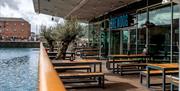 Modern style picnic benches outside BrewDog next to a railing with a large water feature on the other side. Fairylights are hung from the ceiling, wit