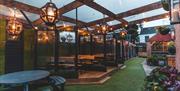 Pubgarden of Larkins Bar. A covered outdoor seating area with wooden beams, large hanging lantern lights, picnic tables, and greenery; some tables are