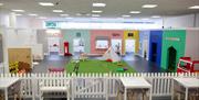 Several playrooms for children around an area with a fake grass in the middle and a small teepee. On the edge of the room is a fence with a seating ar
