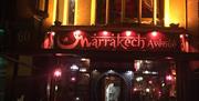 Nighttime view of the entrance to "Marrakech Avenue" restaurant, warmly lit with colorful lanterns, stained glass windows, and decorative signage, cre